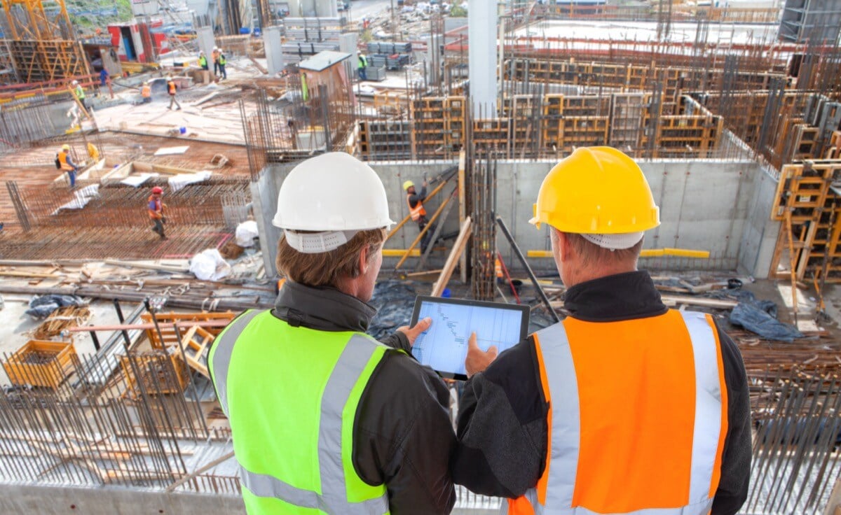 Engineer and architect with hard hats and safety vests on major construction site working on computer tablet; labor shortage productivity concept