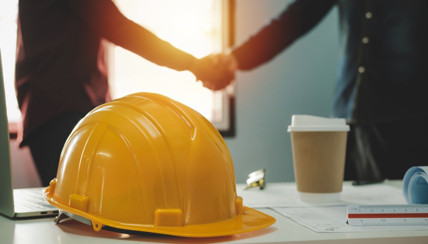 Contractor, with yellow safety helmet on desk, shaking hands with construction worker; working capital costs concept