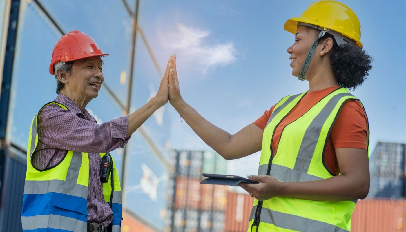 Senior freight forwarder high fives younger worker in the container yard; different generations at work