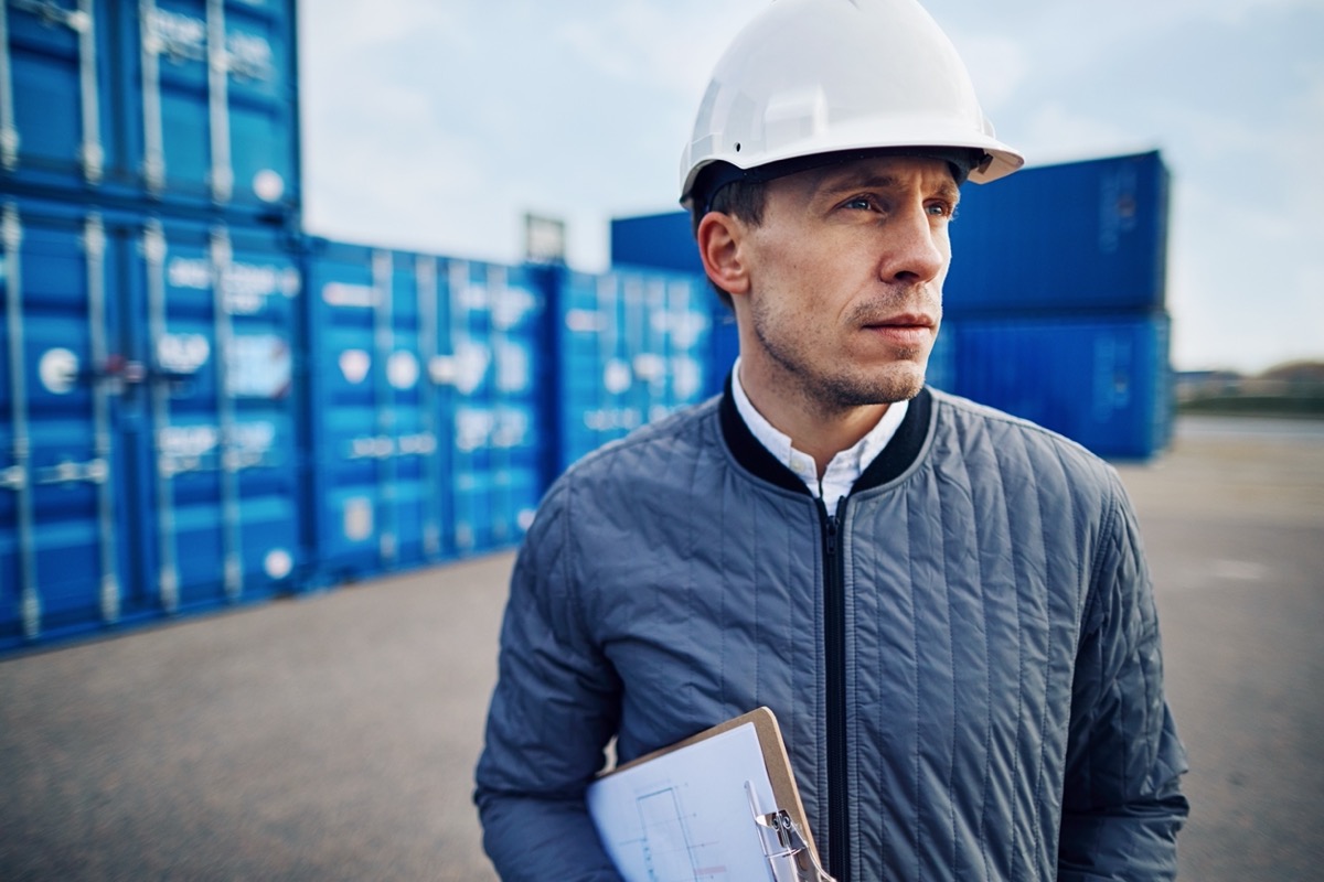 Man wearing a hardhat and holding a clipboard stands on a large commercial shipping dock; paper to software concept