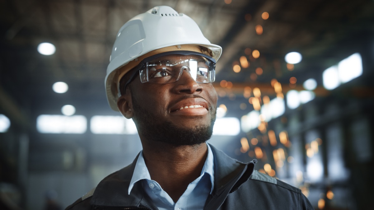 Happy Professional Heavy Industry Engineer Worker Wearing Uniform, Glasses and Hard Hat in a Steel Factory. Smiling African American Industrial Specialist Standing in a Metal Construction Manufacture.