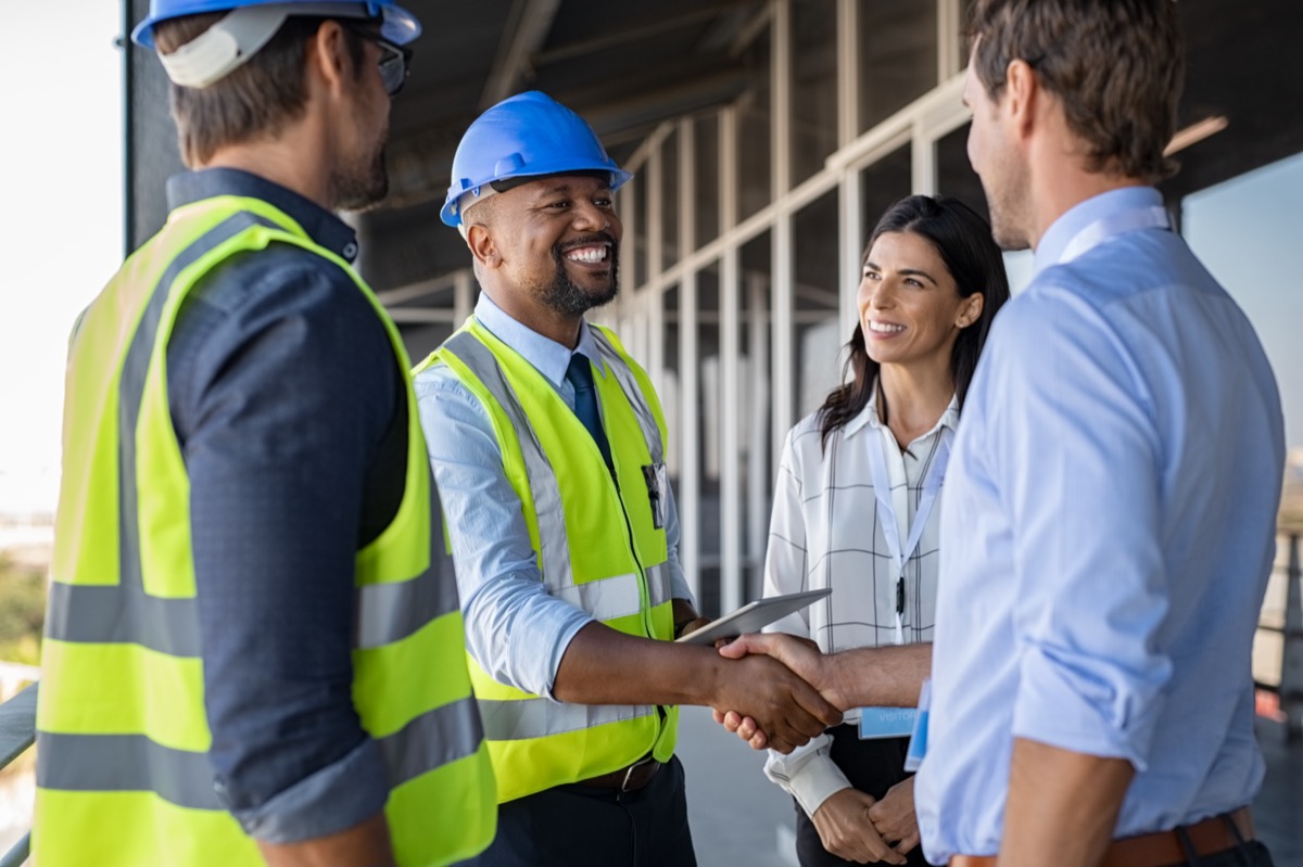 Smiling engineer shaking hands at construction site with businessperson; construction company longevity concept