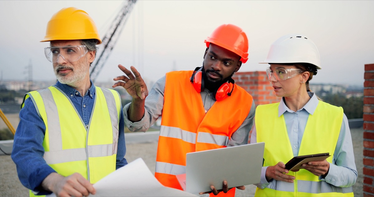 Team of architects and engineers with draft plan of building and laptop computer talking on construction site; construction company longevity concept