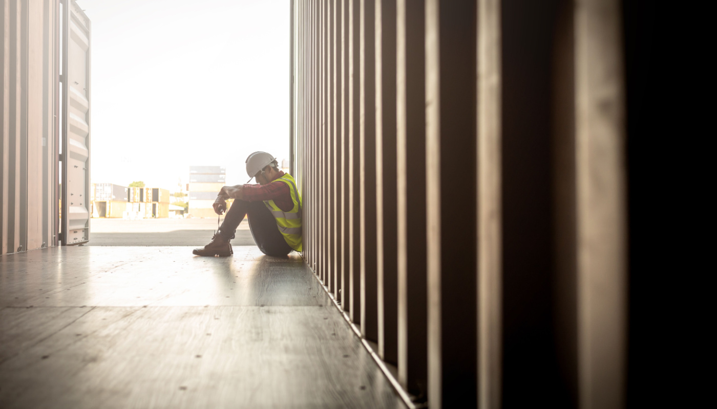 Stressed worker sitting in container box at shipyard; construction industry mental health crisis concept