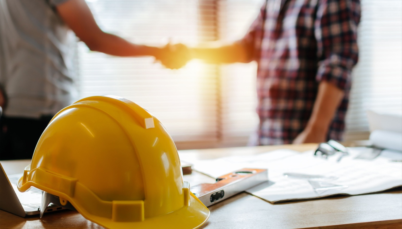 Yellow hard hat on desk, behind which two workers shake hands; create efficiencies concept