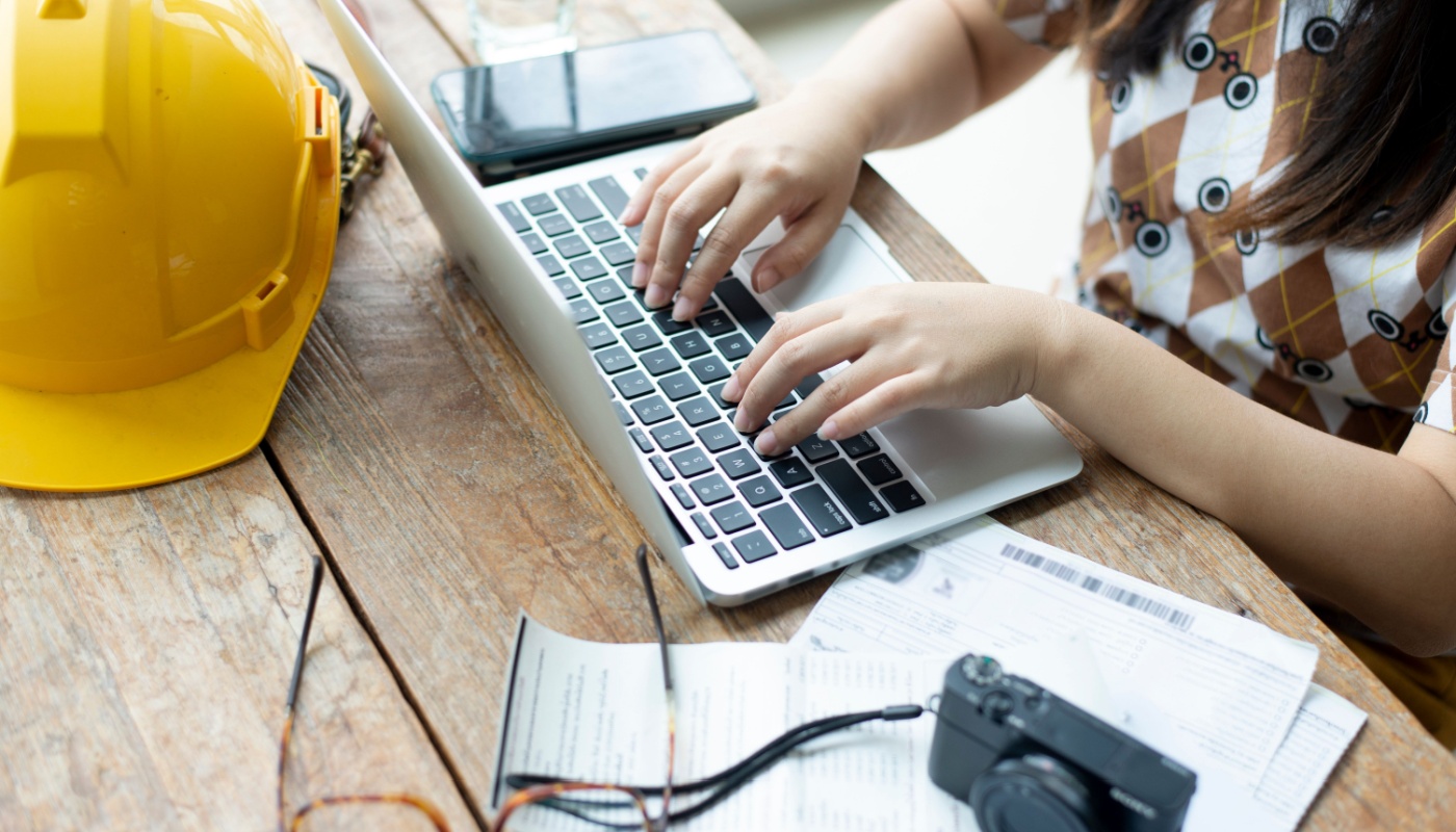 Woman working on laptop with hard hat, phone and camera on the table; create efficiencies concept