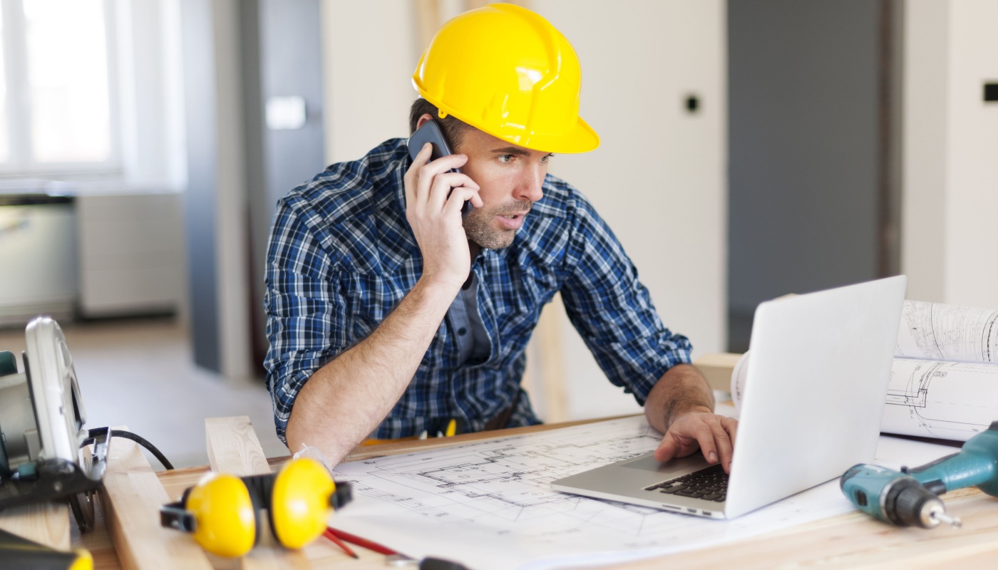 Trade contractor in hard hat at desk, on phone and laptop; create efficiencies concept